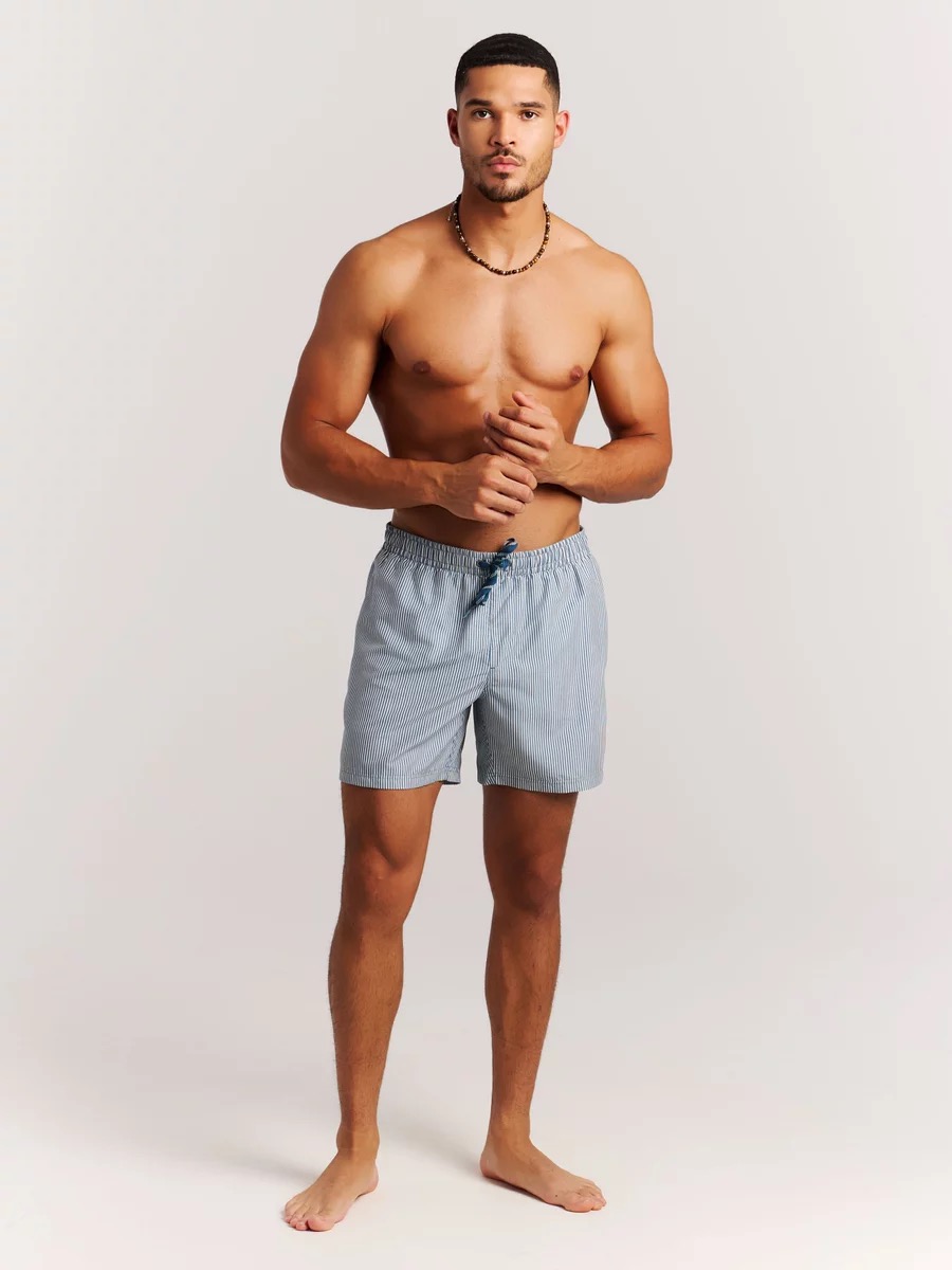 Ein Mann mit dunklem Haar und Halskette, bekleidet mit gestreiften Badeshorts, steht barfuß vor einem hellen Hintergrund.