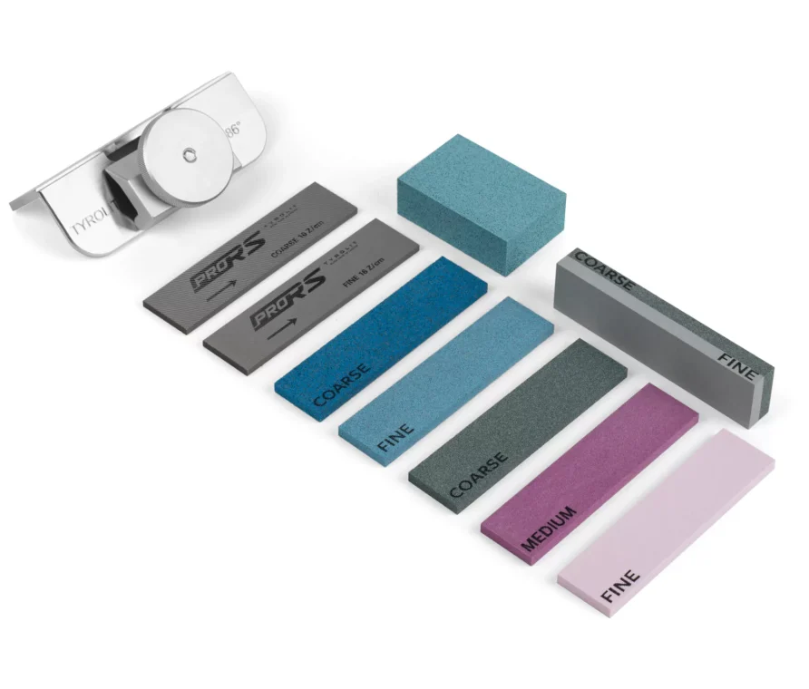 Verschiedenfarbige Schleifsteine mit Beschriftungen wie "Coarse", "Fine", "Medium" und ein Metallwinkel mit Drehknopf liegen auf weißem Grund.