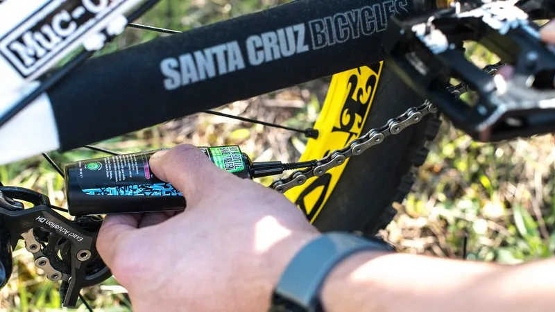 Fahrrad-Kette wird mit Muc-Off Wet Lube geschmiert. Flasche mit grünem Etikett. Fahrrad-Details sichtbar.