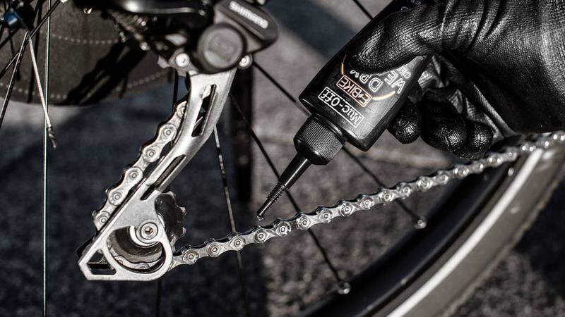 Fahrradkette Ölen Nahaufnahme: Hand ölt mit Muc-Off eBike Kettenöl eine Fahrradkette. Schaltwerk und Reifen sind im Hintergrund.