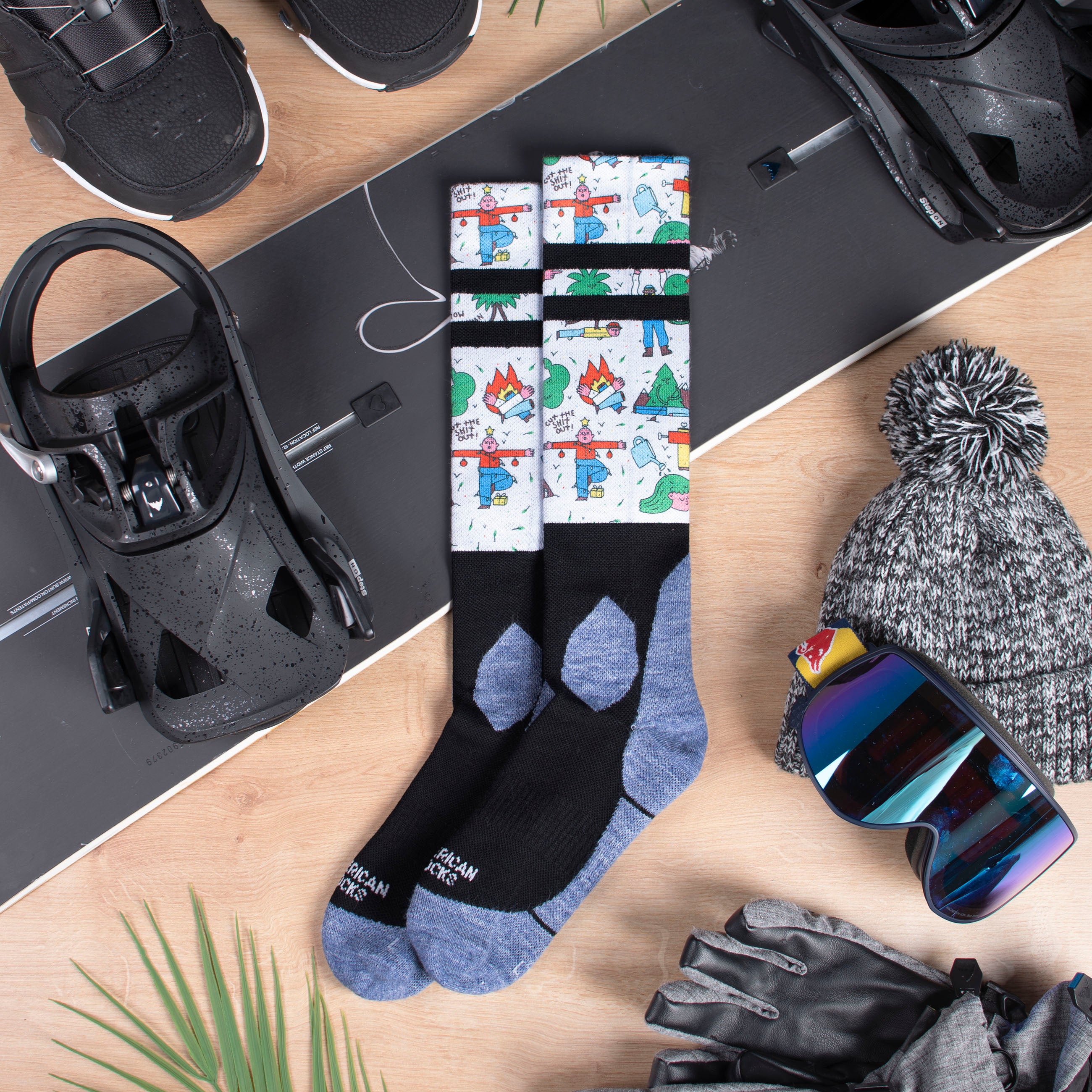 Schneestulpen "Leaf Me Alone" Snow Socks "Leaf Me Alone" auf Snowboard mit Boots, Mütze, Handschuhe und Skibrille auf Holzuntergrund. Socken mit Comic Motiven.