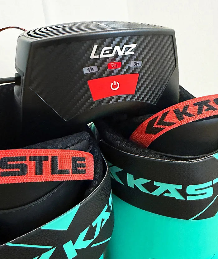 Close-up of ski boots with Lenz dryer. Red straps with black lettering "Kästle" are visible. The dryer has control buttons.