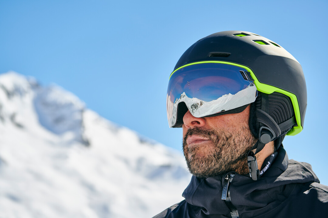 Mann mit Bart, Skihelm und reflektierender Brille. Im Hintergrund verschwommene, schneebedeckte Berge und blauer Himmel.