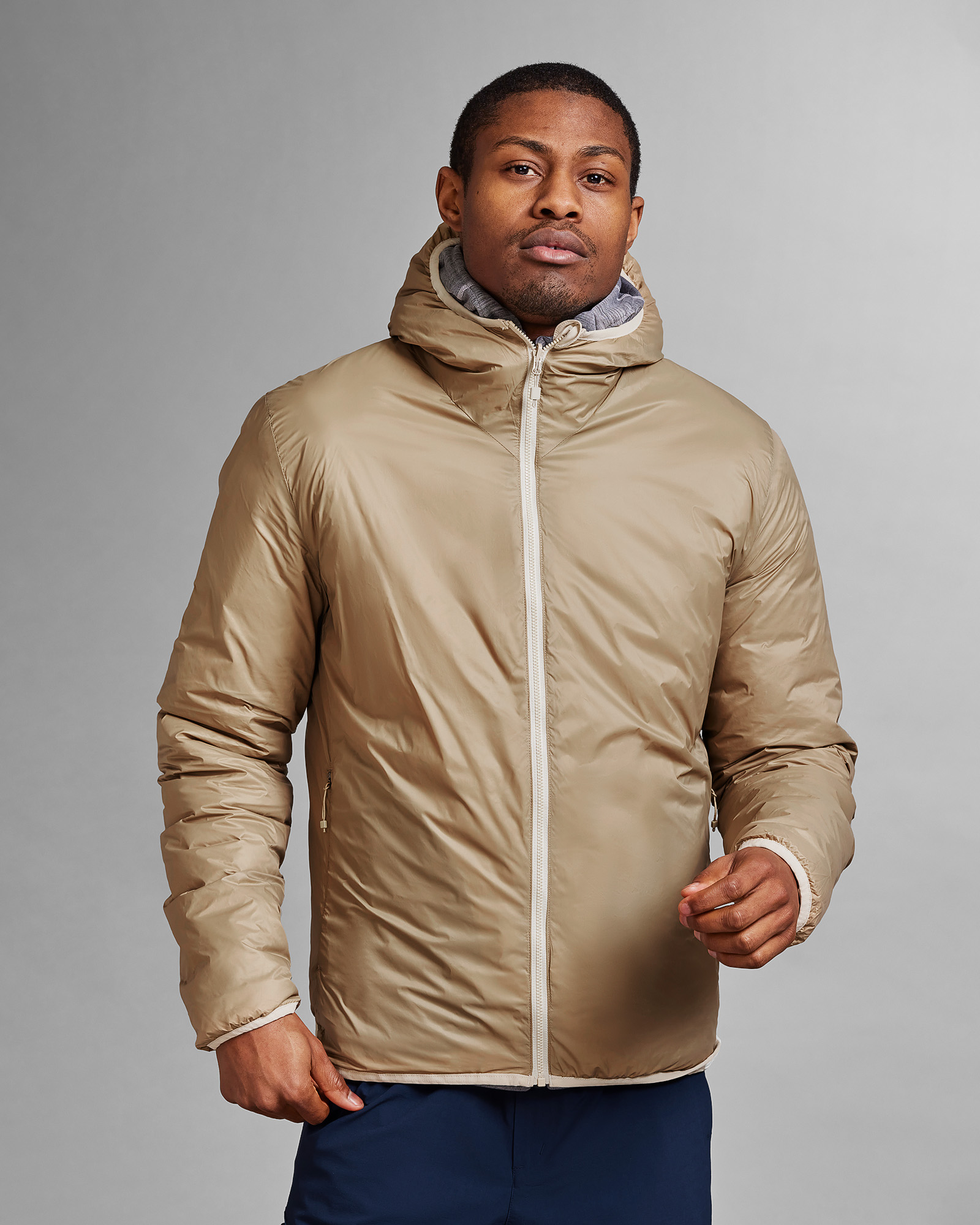 D-3L Wendeweste Sand Ein Mann trägt die D-3L Reversible Light Hood Jacket in Castle Wall Sand. Die Jacke hat eine Kapuze und einen weißen Reißverschluss.