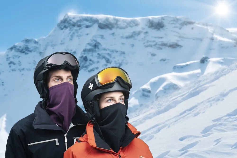 Zwei Personen mit Skihelmen und HeiQ Filter Bandana vor verschneiter Bergkulisse.