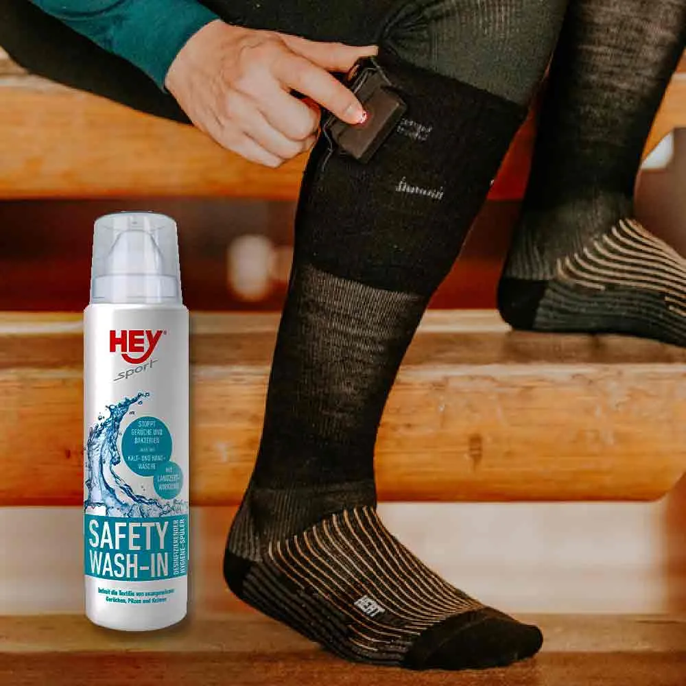 Una persona collega una batteria ricaricabile a un calzino nero riscaldato, accanto al detergente "Hey Sport Safety Wash-In".