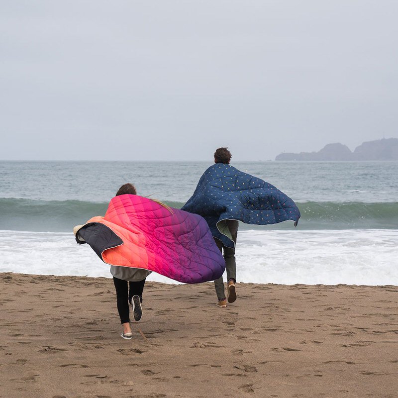 Zwei Personen rennen am Strand, gehüllt in Rumpl Original Puffy Decken; eine in 'Dawn Pixel' mit Farbverlauf, die andere in Blau mit Muster.