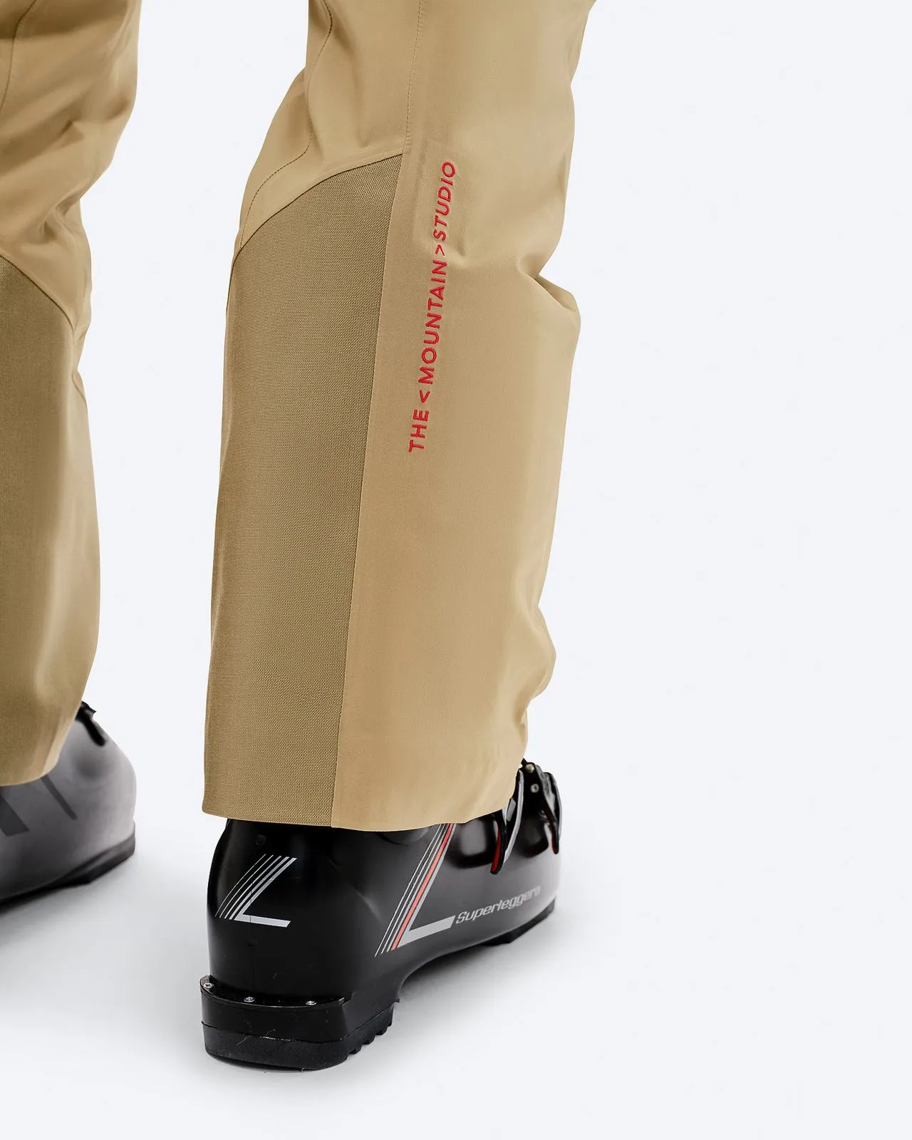 Close-up of the P-1 GORE-TEX trousers in sand color, worn over ski boots. Printed brand logo.