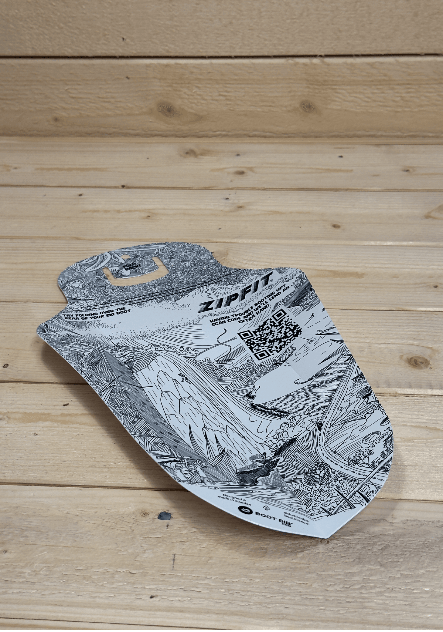 Confezione stampata a forma di scudo, adagiata su uno sfondo di legno chiaro.  La grafica mostra un paesaggio montano abbozzato con codice QR.