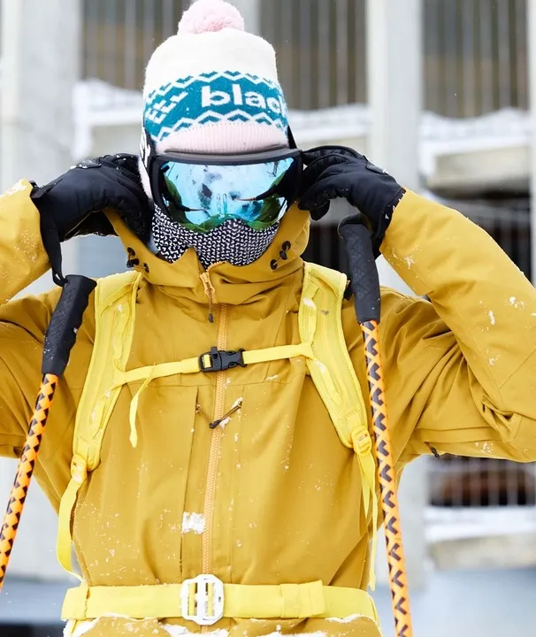 Personne en tenue de ski portant Black Crows Bonnet Crista. Le bonnet est blanc avec un motif bleu et un pompon rose.