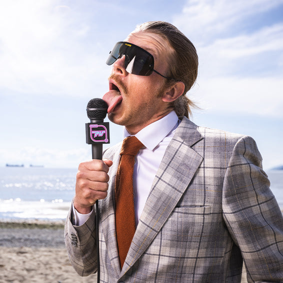 Mann mit Sonnenbrille, Anzug und Krawatte leckt ein Mikrofon am Strand. "Exec" Stil.