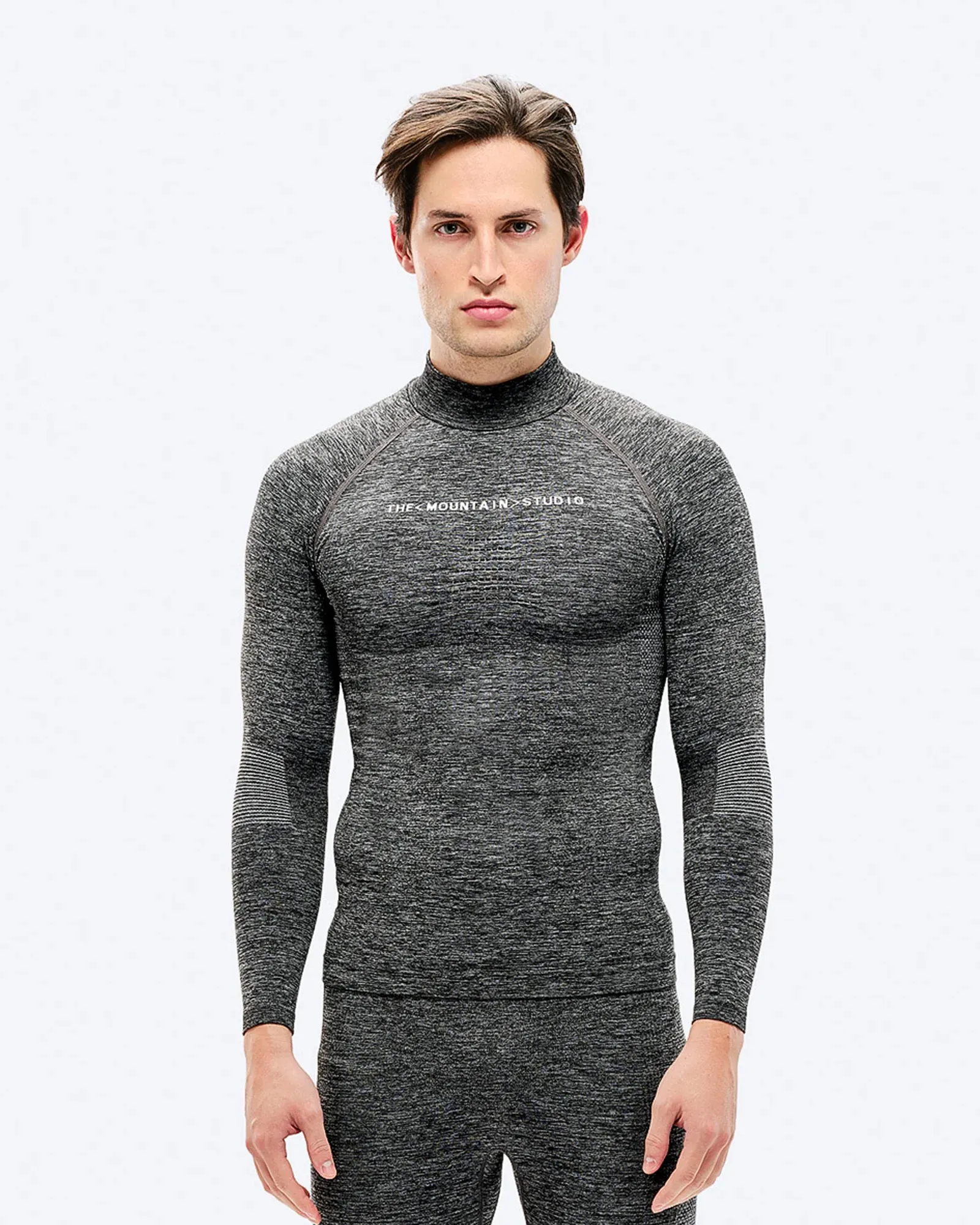 Ein Mann mit braunen Haaren trägt graue Sportbekleidung mit langärmeligem Oberteil und Hose vor einem weißen Hintergrund.