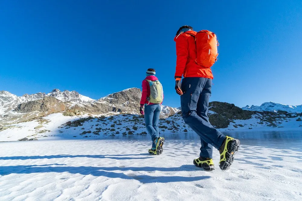 Due persone con artigli da scarpe veloci che camminano sulla neve sullo sfondo di una montagna.