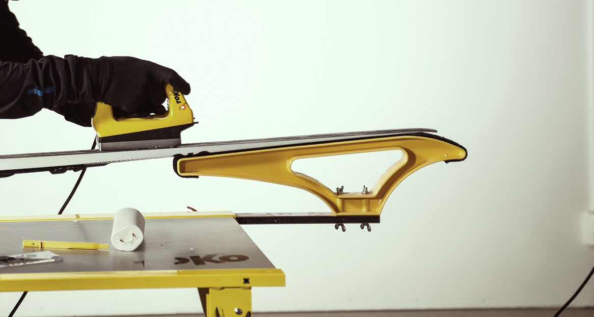 Eine Person wachst einen Ski mit einem gelben Bügeleisen auf einem gelben Skispanner. Ein Tuch und Werkzeuge liegen auf dem Tisch.
