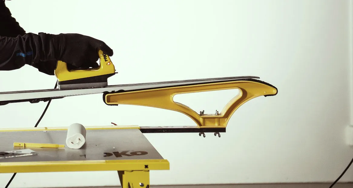 Eine Person wachst einen Ski mit einem gelben Bügeleisen auf einem gelben Skispanner. Ein Tuch und Werkzeuge liegen auf dem Tisch.