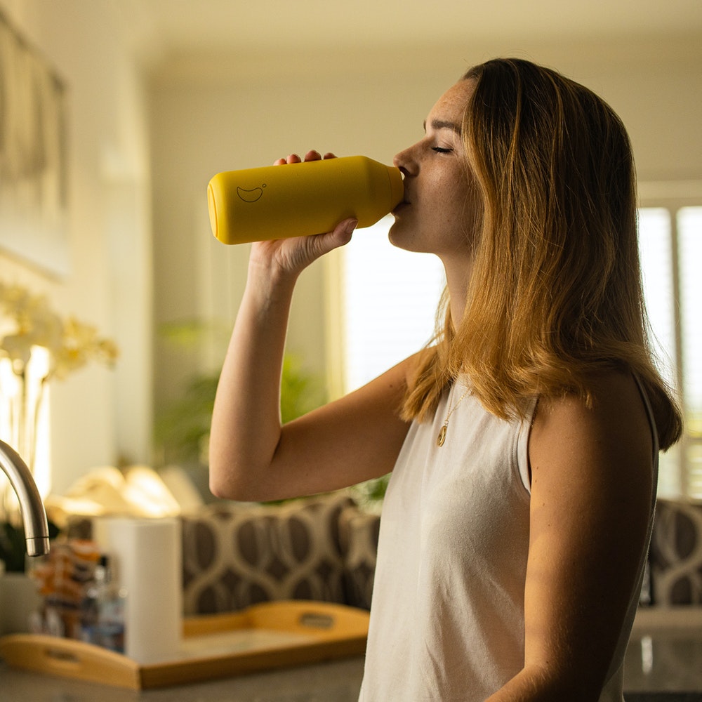 Frau trinkt aus gelber Chillys Trinkflasche Series 2. Flasche im Fokus, heller Hintergrund.