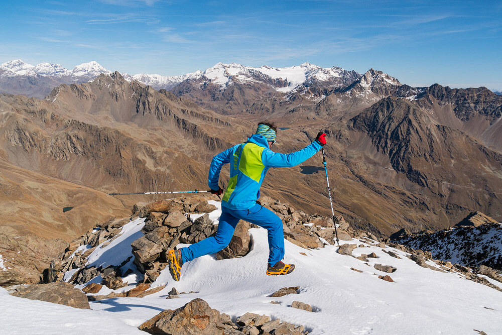 Un homme saute sur une montagne enneigée avec des chaussures de trail running et des griffes de chaussures. En arrière-plan, des montagnes.