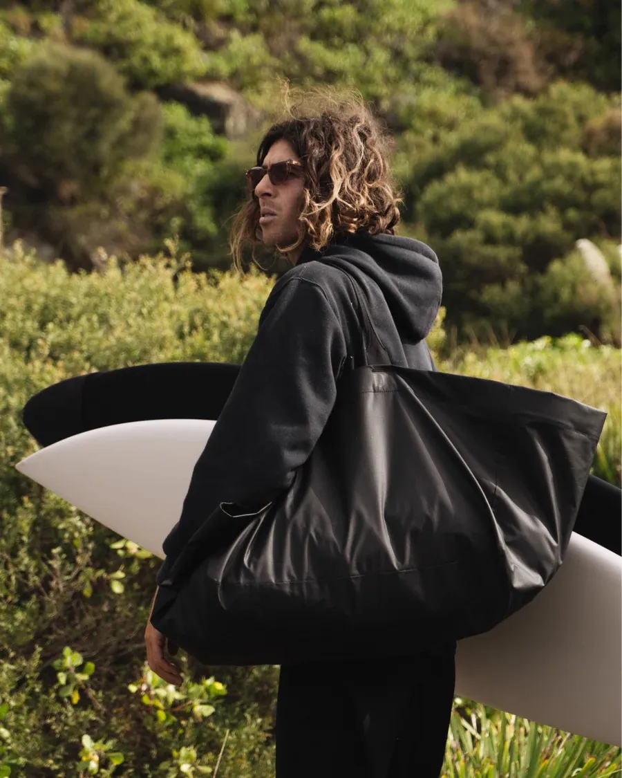 Homme portant une planche de surf et un tote bag noir Essential sur l'épaule, avec une végétation verte en arrière-plan.