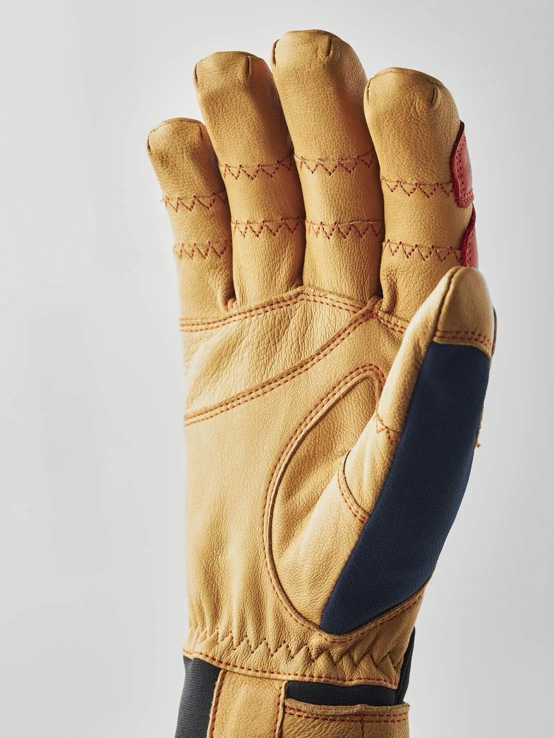 Gros plan sur un gant en cuir marron clair avec une couture rouge et un empiècement bleu foncé au niveau du pouce.
