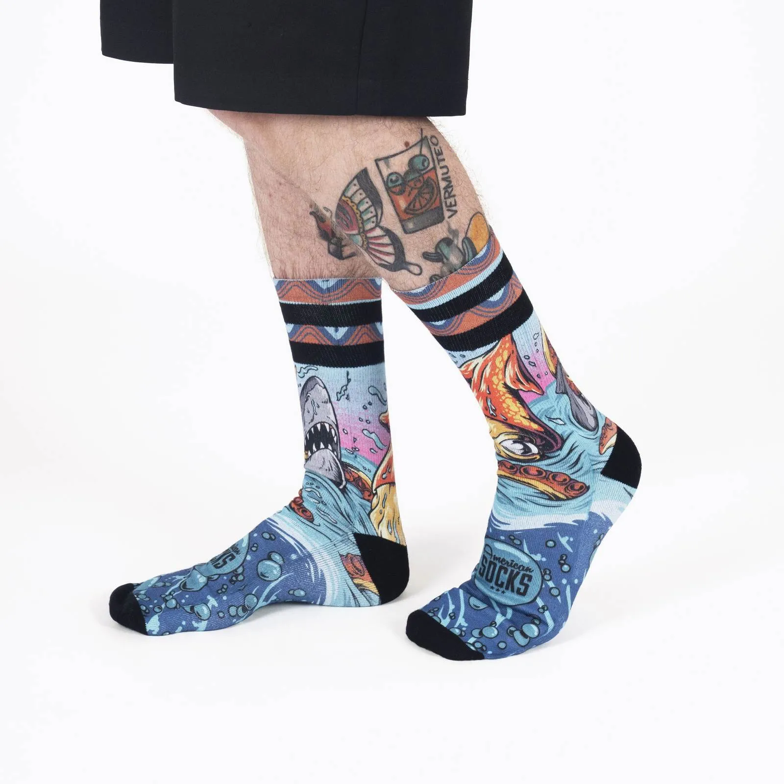 Deux jambes aux mollets tatoués portent des chaussettes bleues `Seamonsters` avec des motifs de requin, de pieuvre et de bulles.
