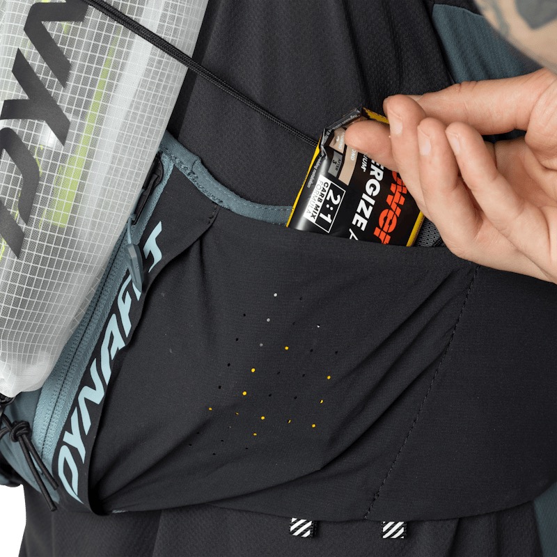 Primo piano di uno zaino da corsa nero Dynafit. Una mano estrae una barretta energetica Powerbar da una tasca laterale. La tasca traforata presenta piccoli punti gialli. È visibile una sacca idrica con motivo a rete bianca.