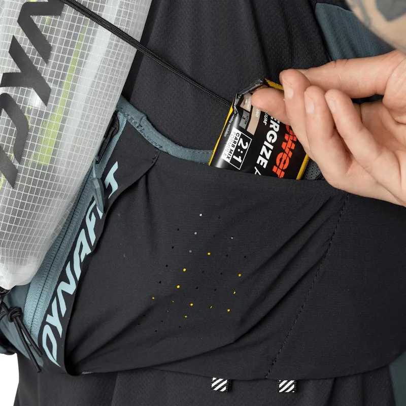 Close-up of a black Dynafit running backpack. A hand is taking a Powerbar energy bar out of a side pocket. The perforated pocket shows small yellow dots. A hydration bladder with a white mesh pattern is visible.