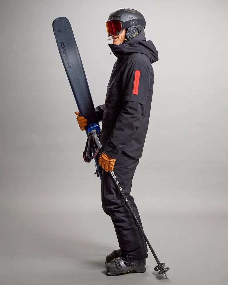 Person in schwarzer Gore Tex Skijacke, Hose, Helm und Skibrille hält Ski und Skistöcke.