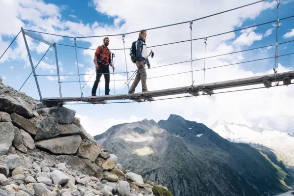 Due escursionisti su un ponte sospeso davanti a un panorama montano sotto un cielo blu con nuvole. Pietre in primo piano.