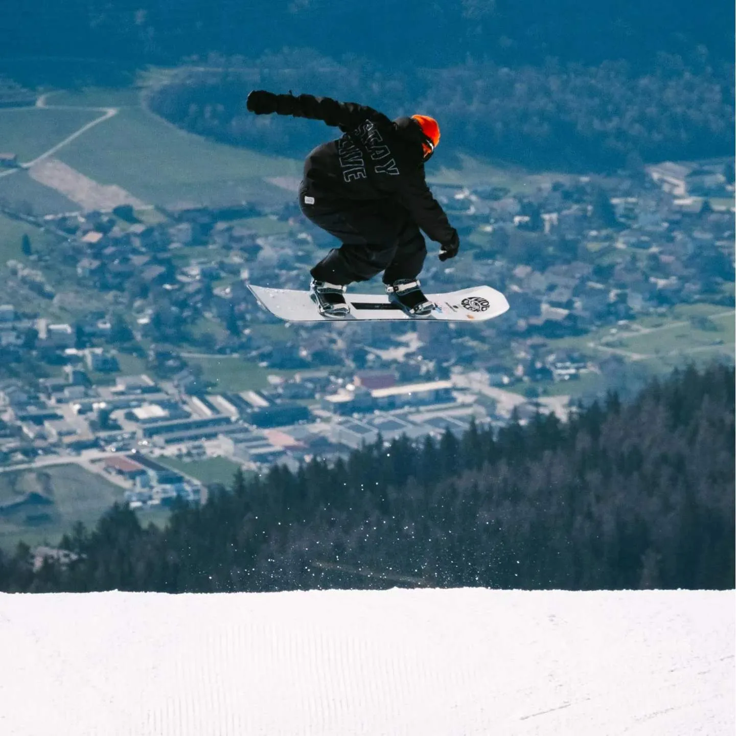 Un snowboarder avec un bonnet orange saute sur une piste préparée sur son snowboard Nidecker Thruster.