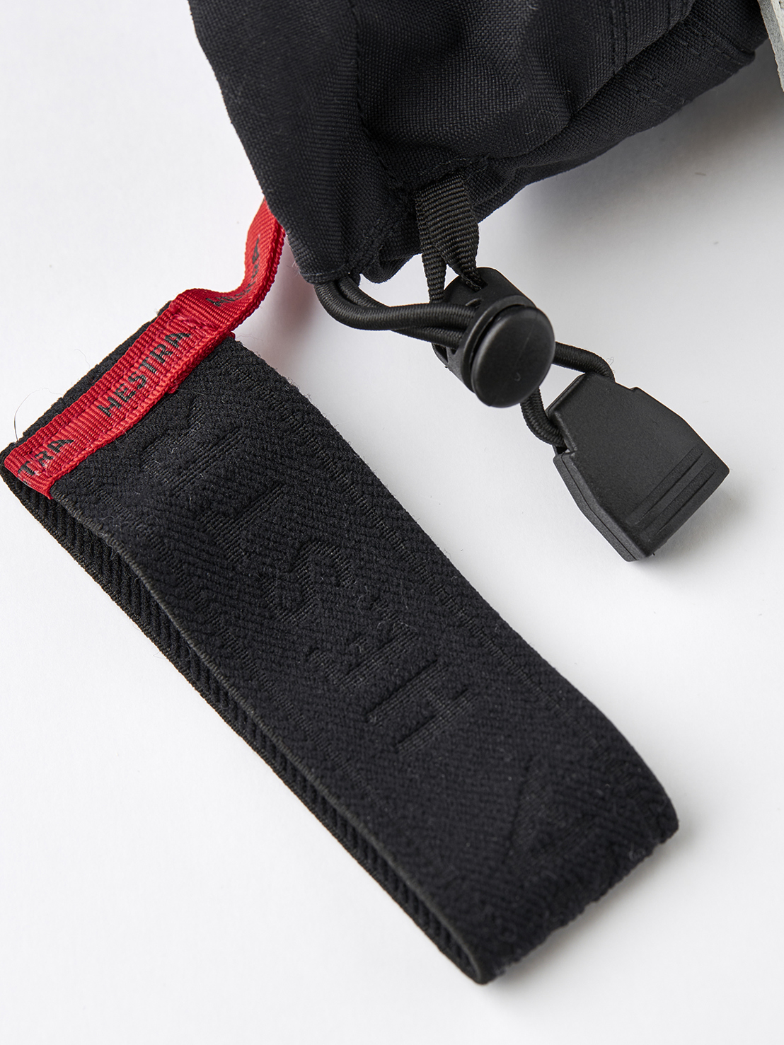 Nahaufnahme eines schwarzen Handschuhs mit roter Schlaufe und schwarzem Gummizug mit Verschluss vor weißem Hintergrund.