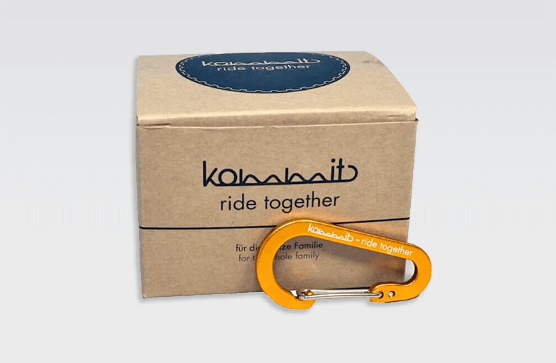 Kommit Karabiner Orange vor einem Karton mit Aufdruck "ride together".