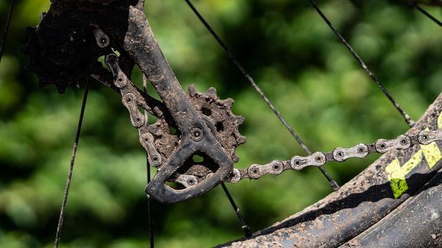 Nahaufnahme einer verschmutzten Fahrradkette mit Ritzel und Schaltwerk vor grünem Hintergrund.
