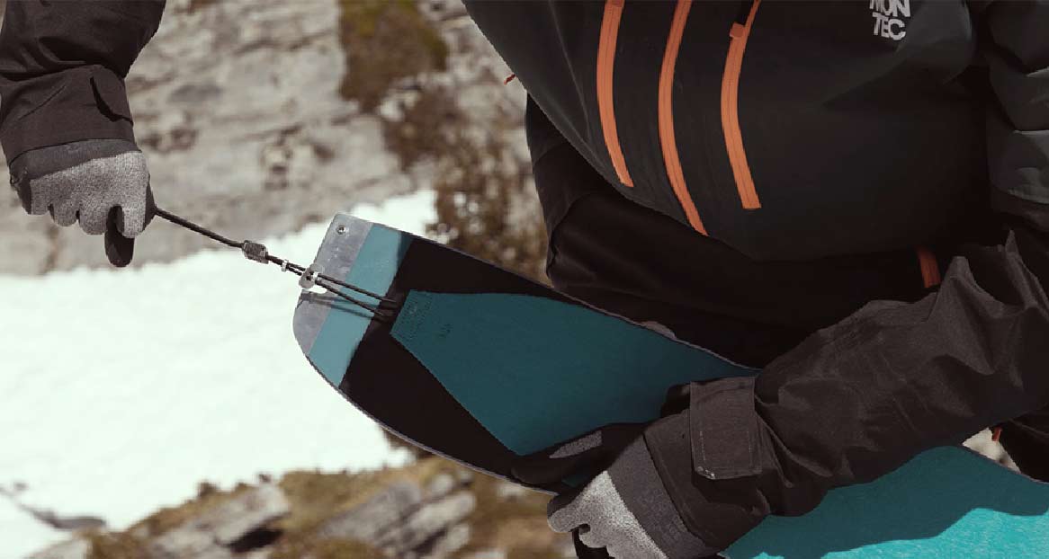 Une personne manipule des peaux de splitboard. Les peaux sont turquoises et noires et sont reliées par une ficelle. Neige en arrière-plan.

