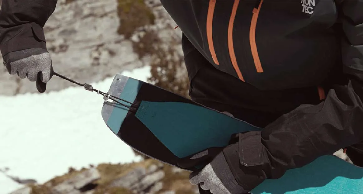 Eine Person hantiert mit Splitboard-Fellen. Die Felle sind türkis und schwarz und mit einer Schnur verbunden. Schnee im Hintergrund.