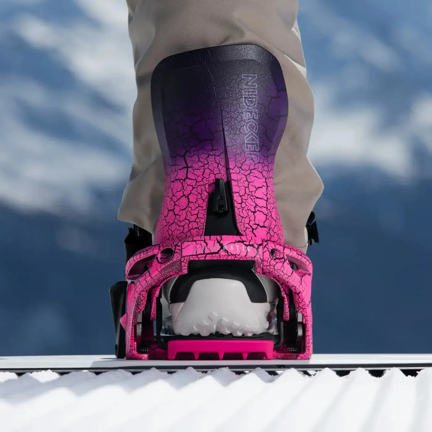 Primo piano di un attacco da snowboard di colore rosa con crepe su uno snowboard su uno sfondo blu sfocato.