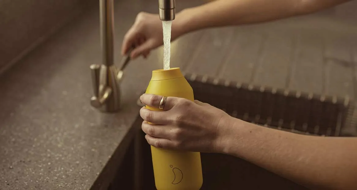 A yellow drinking bottle is filled with water under a tap in a sink. The bottle has a drop-shaped logo.