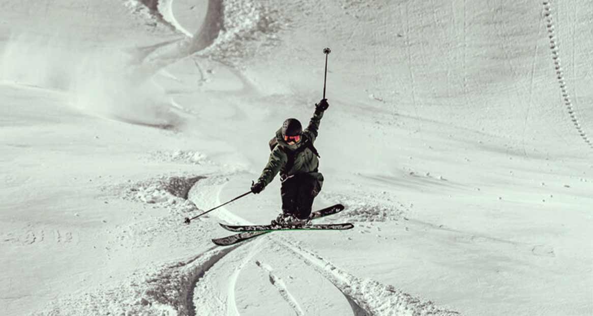 Ein Skifahrer fährt in steilem Gelände mit einer Hand mit einem Stock hoch in der Luft.
