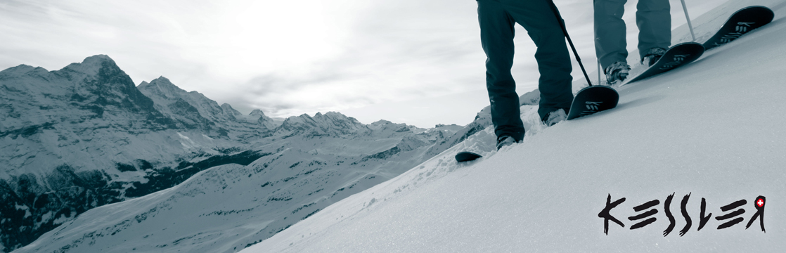 Zwei Skifahrer auf einem schneebedeckten Hang mit einer Bergkette im Hintergrund. Unten rechts ist das Logo "Kessler" zu sehen.