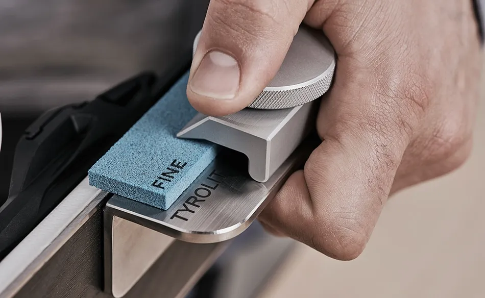 A hand guides the edge of a metal object with a tool and a blue grinding stone "FINE", labeled "Tyrolit".