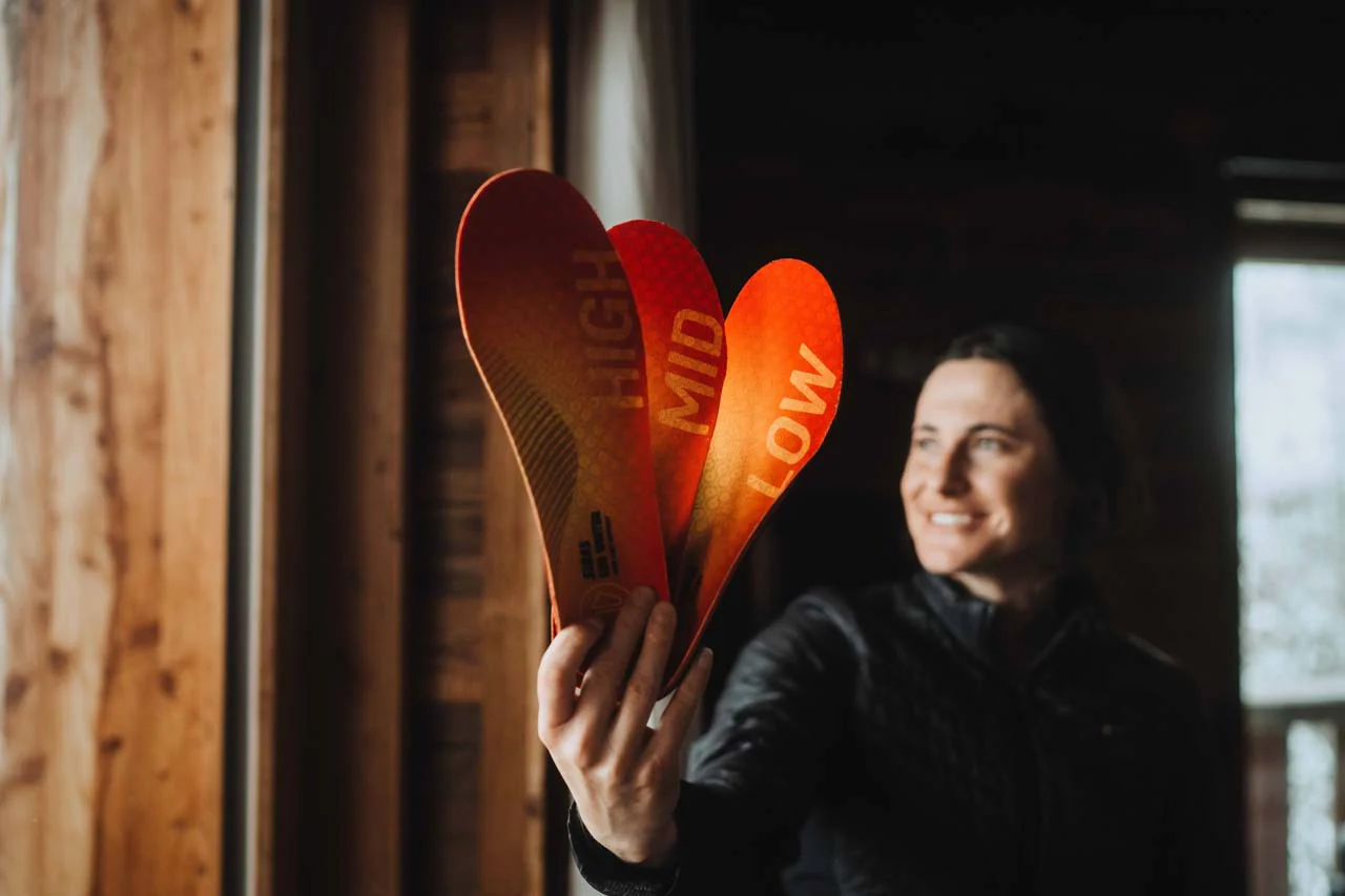 Semelles de ski 3Feet® Eco : Confort écologique pour les skieurs Une femme montre à la caméra trois semelles intérieures rouges et jaunes portant les inscriptions "HIGH", "MID" et "LOW".