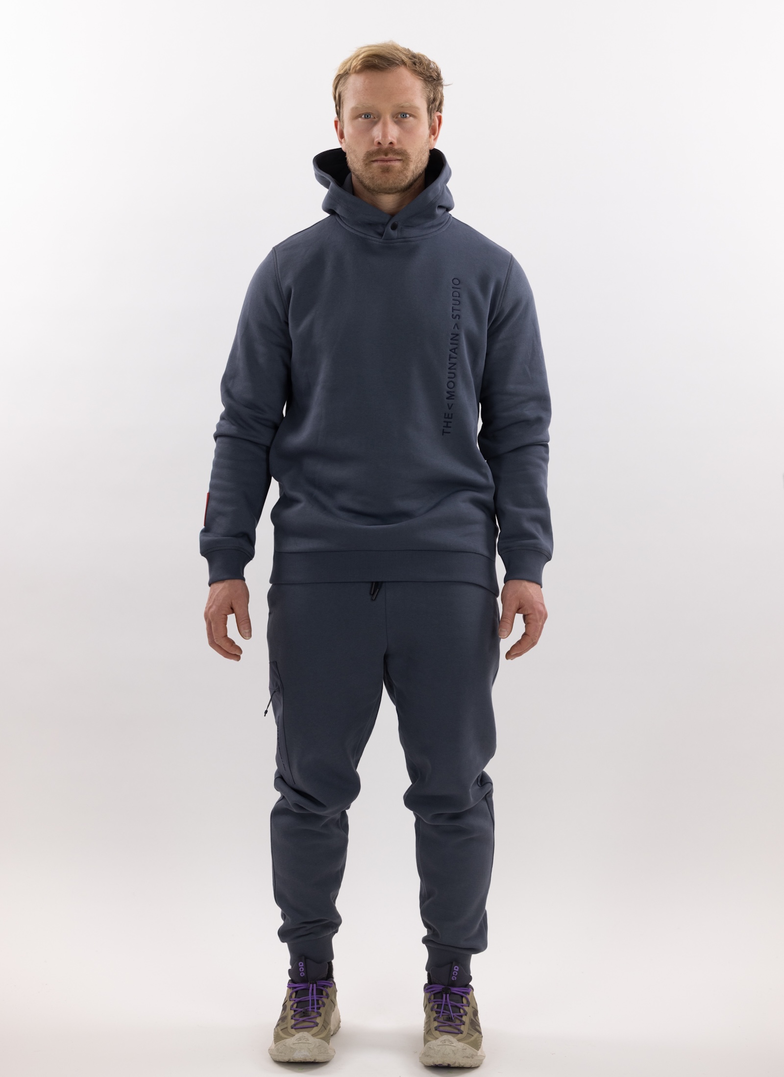 Person in beige 3L Hybrid Shell Jacket, green pants, gloves and ski goggles in front of a neutral background.