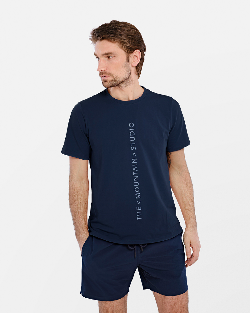Mann trägt dunkelblaues T-Shirt mit vertikalem "The Mountain Studio"-Logo und passende Shorts.