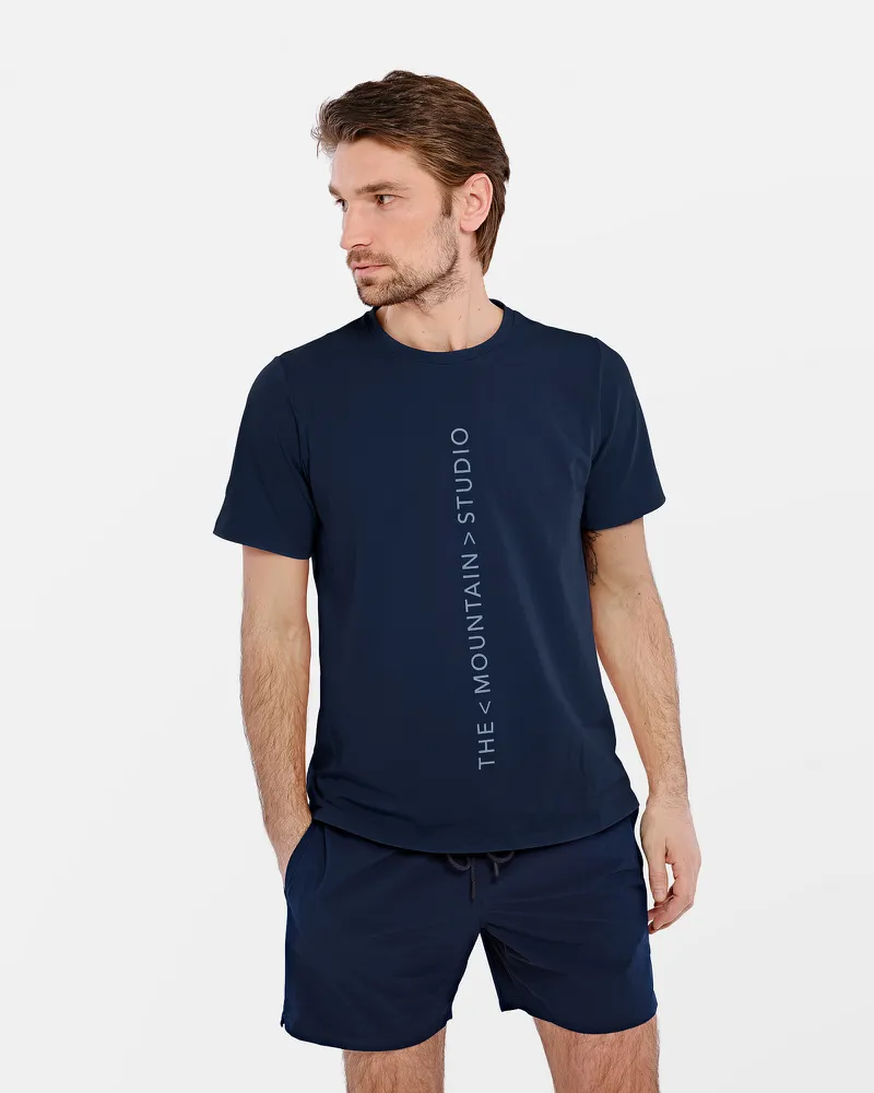 Mann trägt dunkelblaues T-Shirt mit vertikalem "The Mountain Studio"-Logo und passende Shorts.
