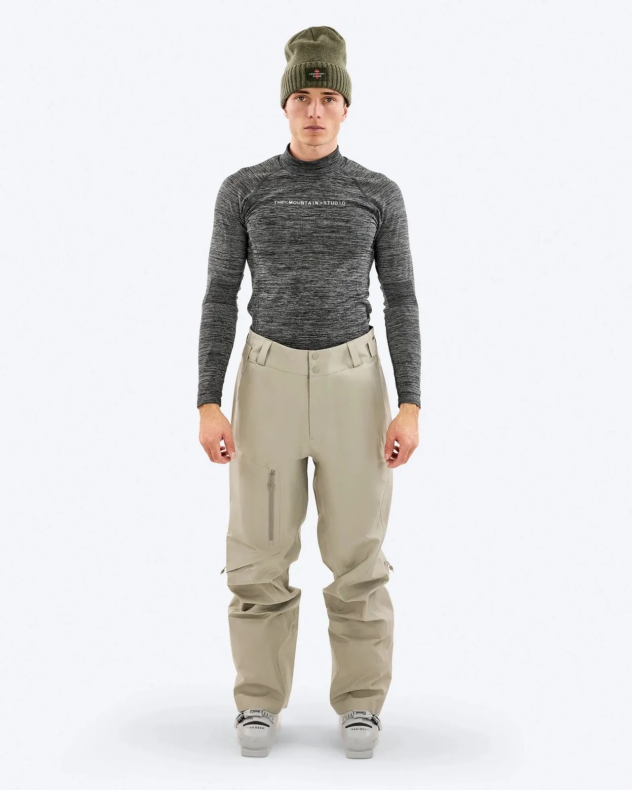 Ein Mann mit olivgrüner Mütze, einem graumelierten Rollkragenpullover, beigen Skihosen und Skischuhen steht vor einem weißen Hintergrund.