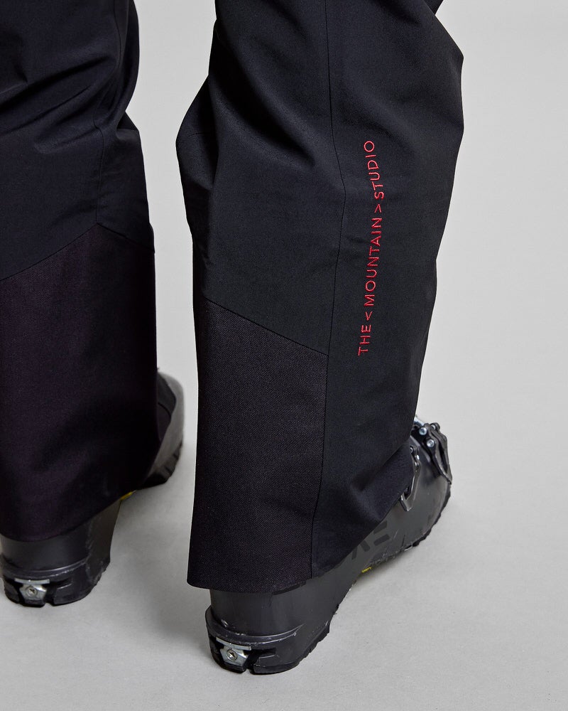 Y-1 HD GORE-TEX 3L Hose Detailansicht der schwarzen Y-1 HD GORE-TEX Pro 3L Shell Pant mit rotem "THE MOUNTAIN>STUDIO" Schriftzug und Skischuhen.