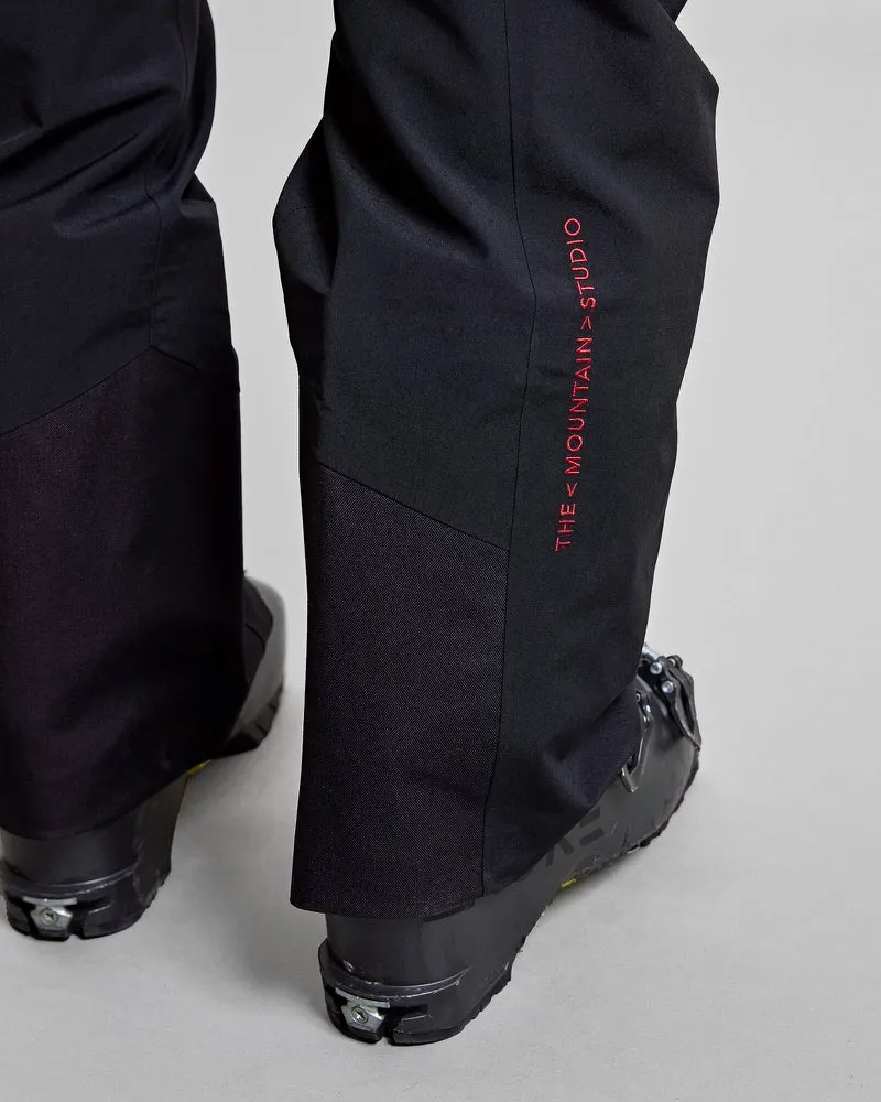 Vue détaillée du pantalon noir Y-1 HD GORE-TEX Pro 3L Shell avec l'inscription rouge "THE MOUNTAIN>STUDIO" et des chaussures de ski.