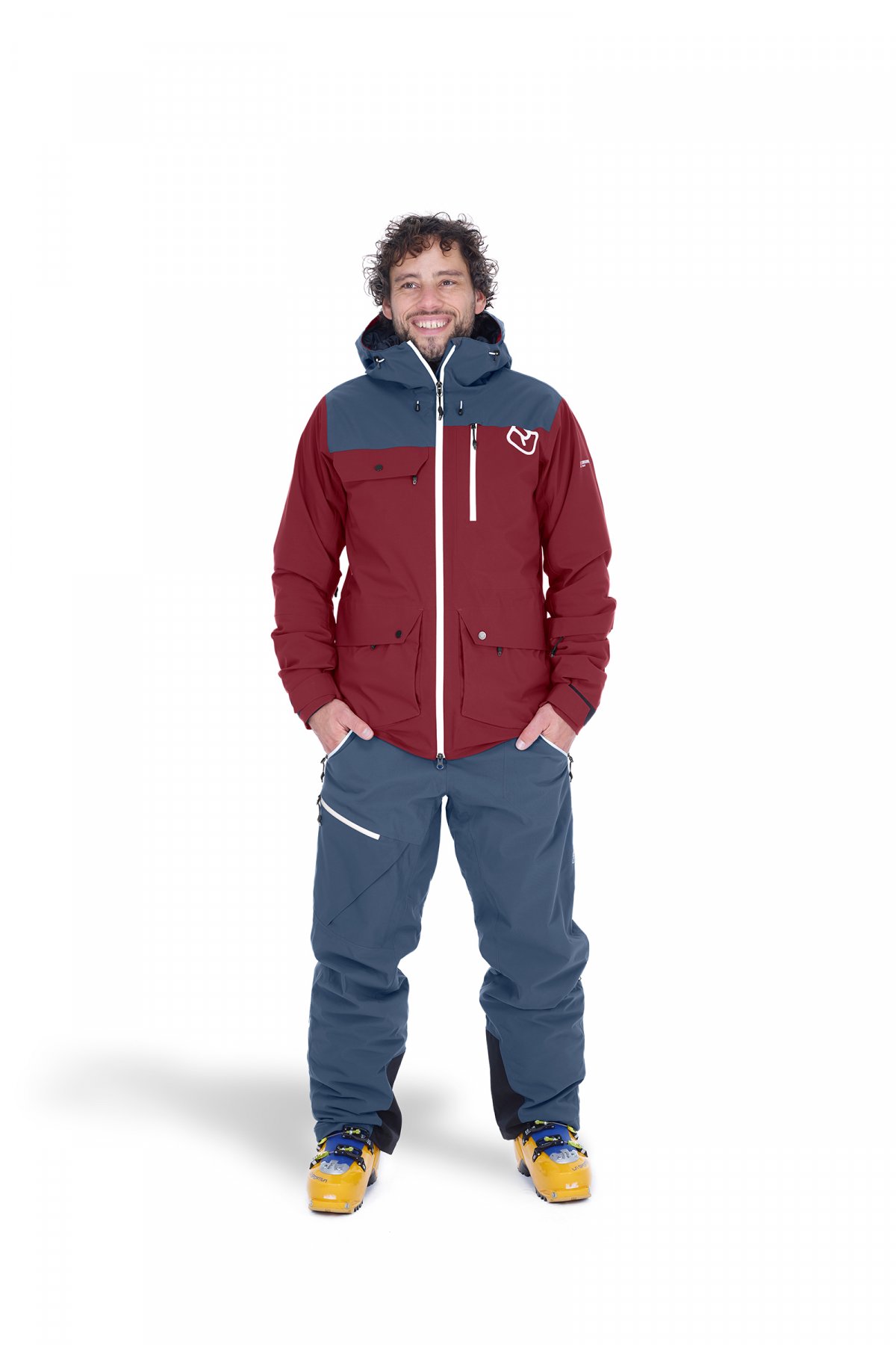 Homme en équipement de ski Ortovox Un homme en Ortovox Andermatt veste et pantalon en Black Steel avec des chaussures de ski, sourit. La veste est rouge et grise.