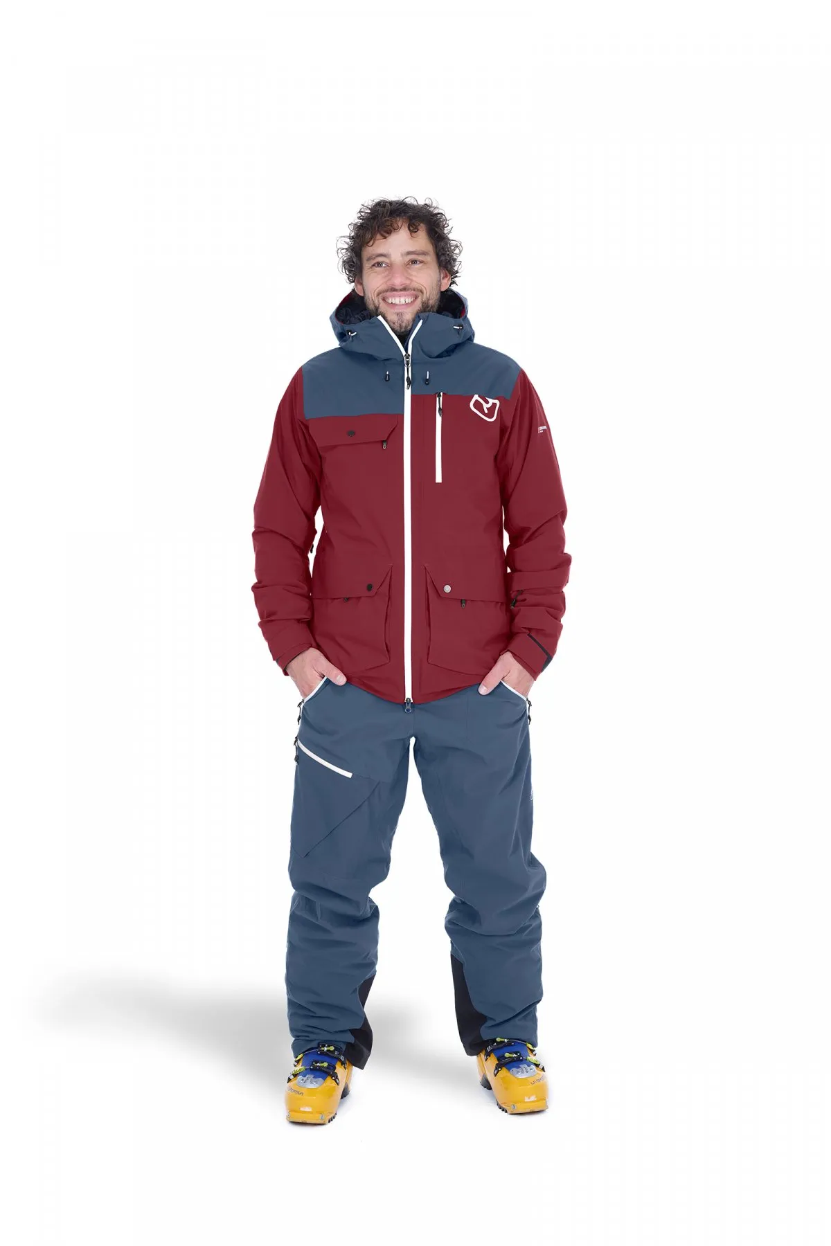 Ein Mann in Ortovox Andermatt Jacke und Hose in Black Steel mit Skischuhen, lächelt. Die Jacke ist rot und grau.