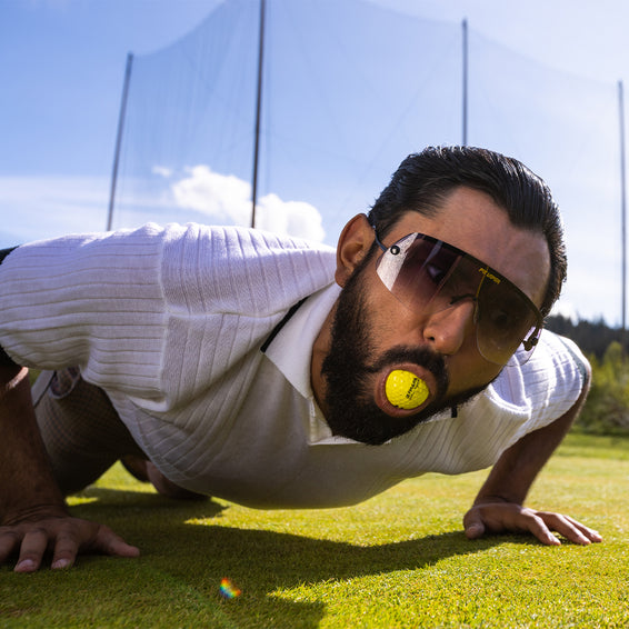 Mann in Liegestützposition mit gelbem Golfball im Mund und Sonnenbrille The Exec.