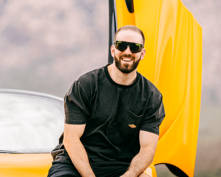 Homme devant une voiture jaune Un homme avec une barbe, un T-shirt noir et des lunettes de soleil pose devant une voiture jaune.